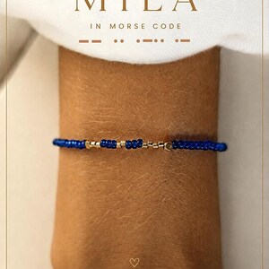 Peut inclure: Un bracelet de perles composé d'une séquence de petites perles rondes bleu foncé alternées avec des perles cylindriques dorées métalliques formant le nom MILA en code Morse. Le texte MILA IN MORSE CODE est imprimé au-dessus.