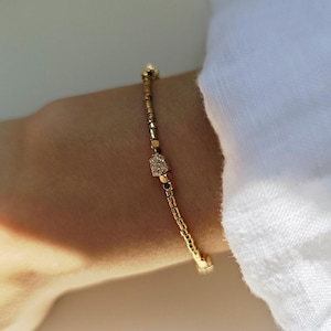 May include: A delicate gold-toned beaded bracelet featuring a small, rectangular charm encrusted with tiny clear stones. The bracelet is composed of uniform metallic cylindrical beads strung together, creating a slender and flexible chain design.