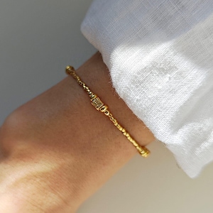 Könnte beinhalten: Ein goldfarbenes Armband mit Perlenmuster und einem rechteckigen Mittelakzent. Das Armband wird am Handgelenk getragen, mit einem weißen Leinenhemd im Hintergrund. Der Schmuck ist schlicht und elegant.