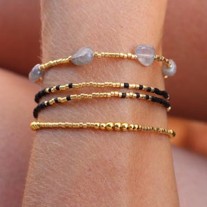 Peut inclure: Un ensemble de quatre bracelets de perles délicats. Le bracelet supérieur présente des perles de rocaille dorées entrecoupées de petites pierres grises translucides et lisses. Deux bracelets centraux sont en perles noires et le bas est doré.