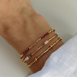 Peut inclure: Un ensemble de trois bracelets de perles délicats composés de perles métalliques dorées, de petites pierres facettées brun rougeâtre foncé et de perles cylindriques lisses de couleur crème, disposés en un design élastique fin et souple.
