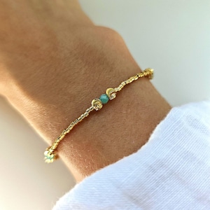 Peut inclure: Un bracelet délicat composé d'un rang continu de petites perles dorées métalliques. Le design est centré sur une unique petite perle ronde de couleur turquoise, encadrée par deux perles sphériques dorées légèrement plus grandes et texturées.