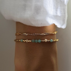 Peut inclure: Deux bracelets portés ensemble. Le bracelet supérieur est une fine chaîne torsadée dorée. Le bracelet inférieur est composé de petites perles dorées alternées avec des perles bleu clair à facettes et des perles roses pâles.