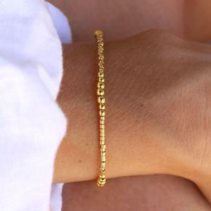 Op de afbeelding: Een delicate armband bestaande uit kleine, gepolijste goudkleurige kralen gerangschikt in een enkele streng. De kralen variëren enigszins in vorm, met een mix van ronde en cilindrische vormen die een textuurrijk en reflecterend oppervlak op de pols creëren.