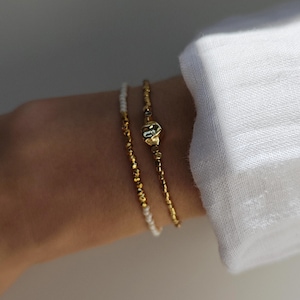 Könnte beinhalten: Zwei goldene Perlenarmbänder und ein weißes Perlenarmband sind an einem Handgelenk zu sehen. Die Armbänder haben ein zartes Design. Die Armbänder werden mit einem weißen Langarmhemd getragen.
