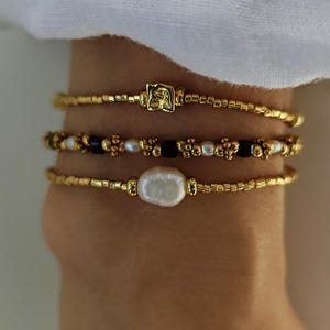 Peut inclure: Trois bracelets de perles avec des perles dorées. Le bracelet supérieur possède une breloque carrée dorée, celui du milieu alterne des perles noires et des perles blanches, et le bracelet inférieur présente une grosse perle blanche irrégulière.