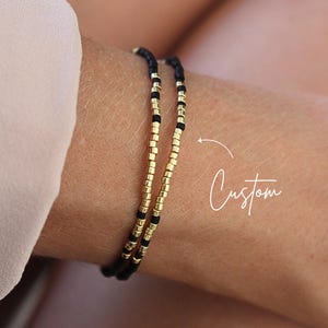 Könnte beinhalten: Zwei zarte Armbänder aus abwechselnd kleinen schwarzen und metallisch goldfarbenen zylindrischen Perlen. Die Armbänder weisen ein wiederkehrendes Muster dieser Perlen auf, wobei ein weißer Pfeil mit dem handschriftlichen Text Custom auf den Schmuck zeigt.