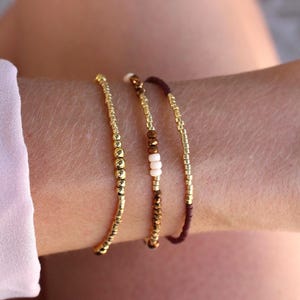 Peut inclure: Un ensemble de trois bracelets de perles délicats. Un bracelet présente des perles dorées métalliques uniformes. Le deuxième combine des perles dorées, bronze facettées et crème mat. Le troisième alterne des perles dorées et marron foncé.