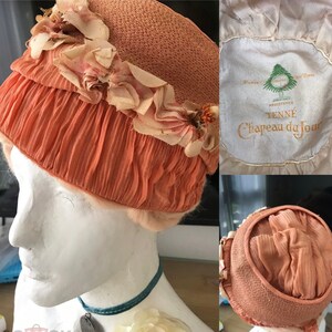 May include: A peach-colored hat with a ruffled trim and floral embellishments. The hat is labeled "TENN&Eacute;  Registered  Chapeau du Jour  Paris  New York".