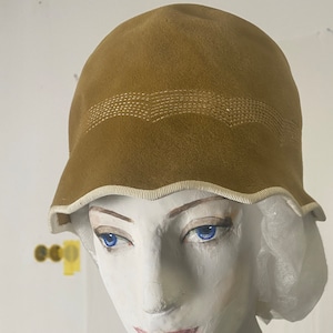 May include: A brown velvet cloche hat with a white trim and a stitched design. The hat is worn by a mannequin with a pearl necklace.