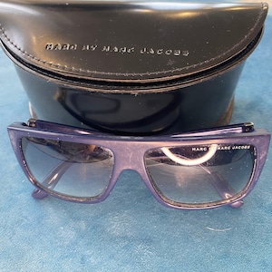 May include: A pair of dark blue sunglasses with a black case. The sunglasses have a rectangular shape and the case has the text "MARC BY MARC JACOBS" on it.
