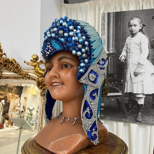 Könnte beinhalten: Ein Schaufensterpuppenkopf mit einem blau-silbernen, perlenbesetzten Kopfschmuck. Der Kopfschmuck hat ein türkisfarbenes Mittelteil und ist mit blauen und silbernen Perlen verziert. Die Mannequin wird auf einem goldfarbenen Sockel präsentiert.