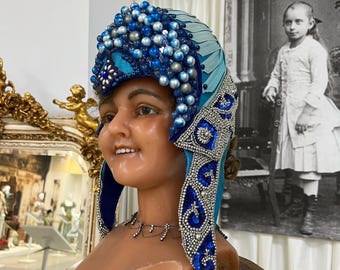 May include: A mannequin head wearing a blue and silver beaded headdress. The headdress features a central turquoise accent and is adorned with various sizes of blue and silver beads. The mannequin is displayed on a gold-colored pedestal.