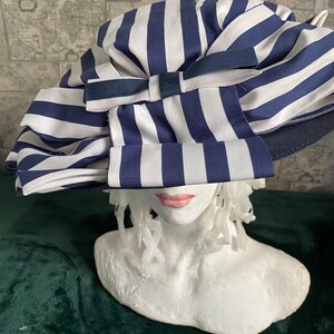 May include: A blue and white striped hat with a large bow. The hat is made of a soft fabric and has a wide brim. The bow is made of a matching blue fabric. The hat is displayed on a white bust.