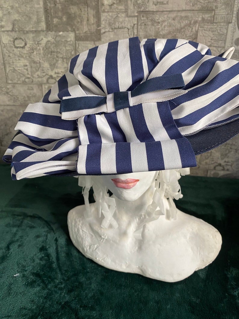 May include: A blue and white striped hat with a large bow. The hat is made of a soft fabric and has a wide brim. The bow is made of a matching blue fabric. The hat is displayed on a white bust.