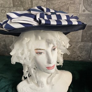 May include: A navy and white striped hat with a wide brim. The hat has a large, layered design with a bow. The hat is displayed on a white mannequin head.