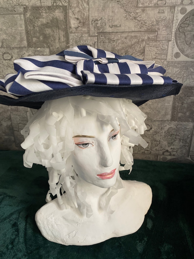 May include: A navy and white striped hat with a wide brim. The hat has a large, layered design with a bow. The hat is displayed on a white mannequin head.