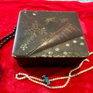 May include: A dark, rectangular box with a decorative lid featuring a bird and floral design. A black beaded necklace with a tassel and a white beaded necklace with a blue accent are next to the box, all on a red surface.