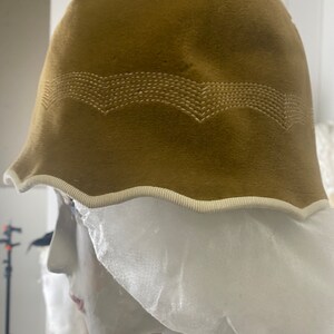 May include: A brown felt cloche hat with a white trim and a white stitched design. The hat is on a white mannequin head.