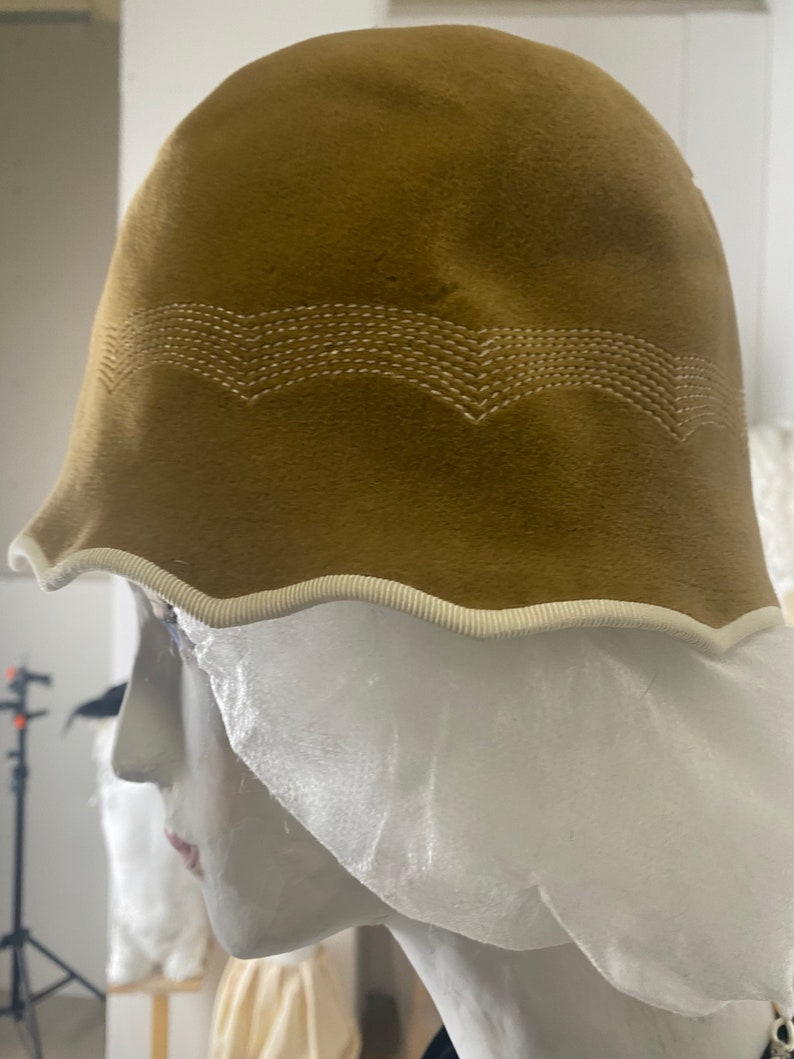 May include: A brown felt cloche hat with a white trim and a white stitched design. The hat is on a white mannequin head.