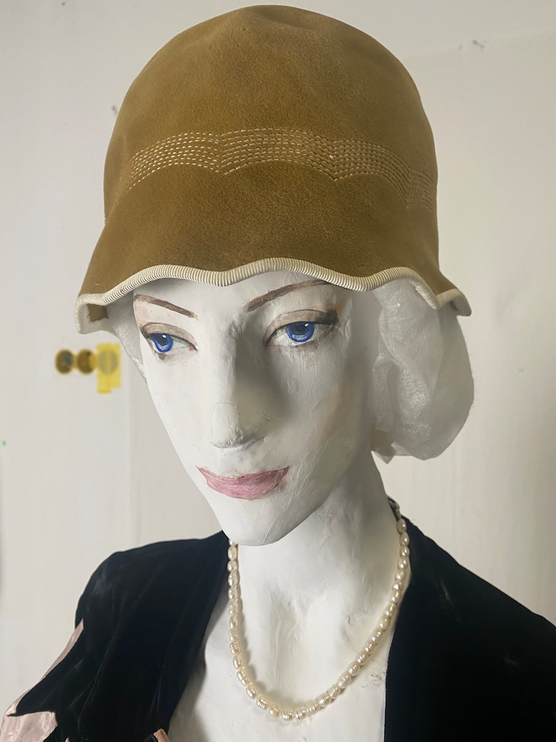 May include: A brown velvet cloche hat with a white trim and a stitched design. The hat is worn by a mannequin with a pearl necklace.