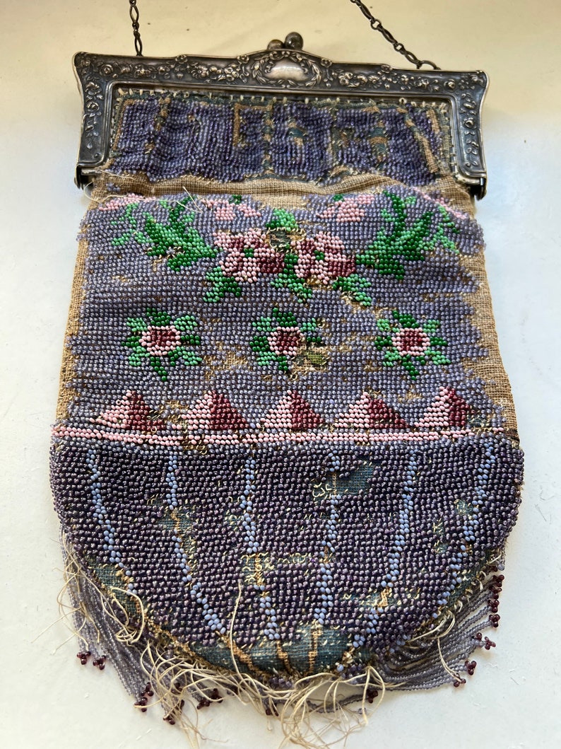 May include: A vintage beaded purse with a silver clasp and floral design. The purse is made of small, purple beads and has a fringe of beads hanging from the bottom.