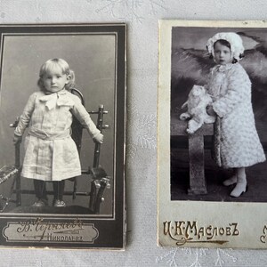 May include: Two antique black and white portrait photographs. One shows a young child in a dress seated on a chair. The other shows a child in a coat holding a stuffed animal. Both photos have text at the bottom.