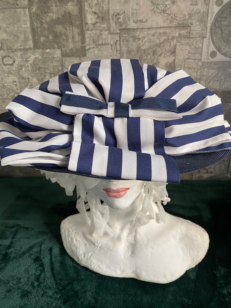 May include: A blue and white striped sun hat with a large bow. The hat is displayed on a white bust. The hat's brim is dark blue.