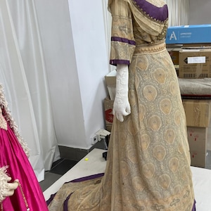 May include: A vintage dress on a mannequin. The dress has a beige and gold pattern with purple trim. The dress has long sleeves and a long skirt with a train. The mannequin is wearing a white wig with feathers. The dress is reminiscent of the Edwardian era.