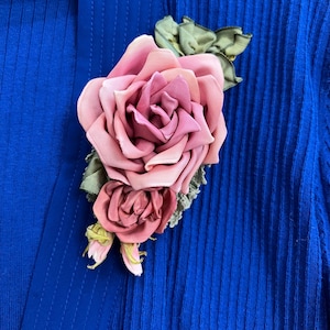 Può includere: Un fermaglio a forma di rosa in tessuto rosa e malva, con foglie verdi e un retro in pizzo. L'accessorio floreale è appuntato su un tessuto blu a coste. Un fermaglio decorativo.
