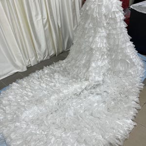 May include: A white, layered skirt with a long train. The skirt is made of multiple tiers of ruffled fabric, creating a voluminous and textured appearance. The garment is displayed on a mannequin.