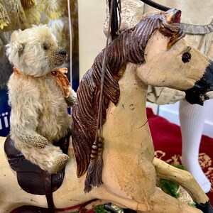 May include: A vintage-style teddy bear with a light brown fur sits atop a wooden carousel horse. The horse is painted in cream and brown tones, with a black muzzle and mane. The bear wears a small orange ribbon around its neck.