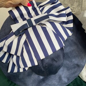 May include: A navy blue sun hat with a wide brim and a large bow. The bow features white and navy blue vertical stripes. A smaller navy blue and white ribbon is tied on top of the bow. The hat is made of woven material.