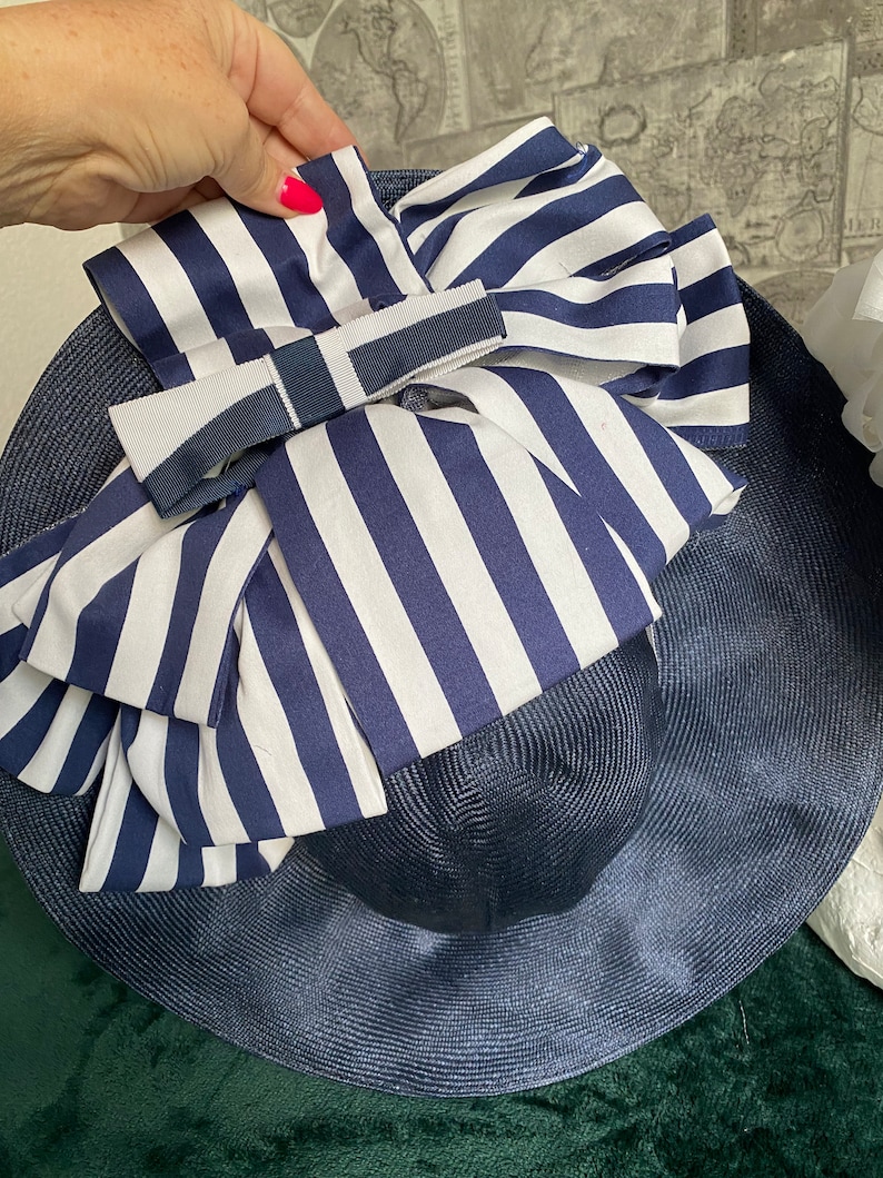 May include: A navy blue sun hat with a wide brim and a large bow. The bow features white and navy blue vertical stripes. A smaller navy blue and white ribbon is tied on top of the bow. The hat is made of woven material.