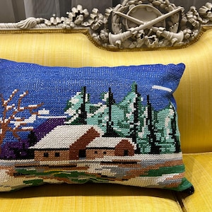 以下が含まれることがあります： 黄色いソファの上に置かれた、青と白の刺繍が施された枕。冬景色が刺繍されており、家、木、雪が描かれている。