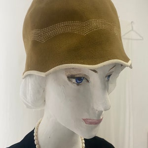 May include: A brown felt cloche hat with a white stitched design around the crown. The hat has a white trim around the brim.