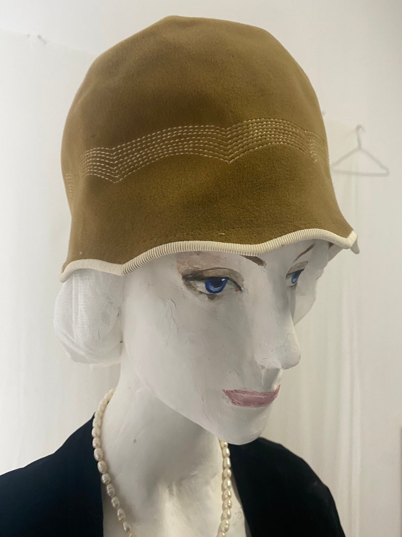 May include: A brown felt cloche hat with a white stitched design around the crown. The hat has a white trim around the brim.