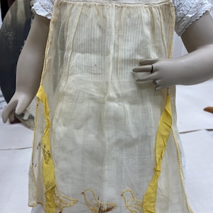 Puede incluir: Un delantal crema de estilo vintage con ribete amarillo y pollitos bordados. El delantal presenta un patrón de rayas verticales y está adornado con una cinta amarilla brillante. La parte superior del delantal tiene tres pollitos bordados, y la inferior tiene tres más.