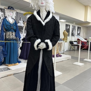 May include: A mannequin wearing a black coat with white fur trim, a long black skirt, and a decorative hat with white and beige accents. The mannequin is part of a display of vintage clothing.