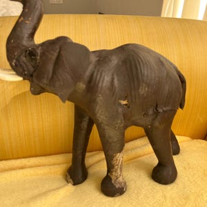 May include: A dark brown elephant figurine with ivory-colored tusks. The elephant has a raised trunk and is standing on four legs. The figurine is placed on a yellow surface.