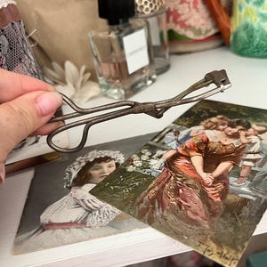 以下が含まれることがあります： ヒンジデザインとエイジングメタル仕上げが施された、ヴィンテージの金属製まつげカーラー。カーラーは、湾曲したハンドルと小さな長方形のパッドを備えています。画像には、アンティークのポストカードと香水瓶も写っています。