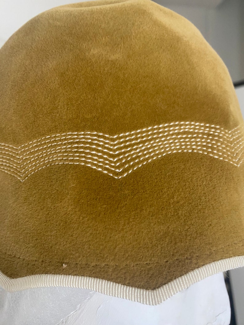 May include: A brown felt hat with a white stitched design around the brim. The hat has a white trim around the edge.