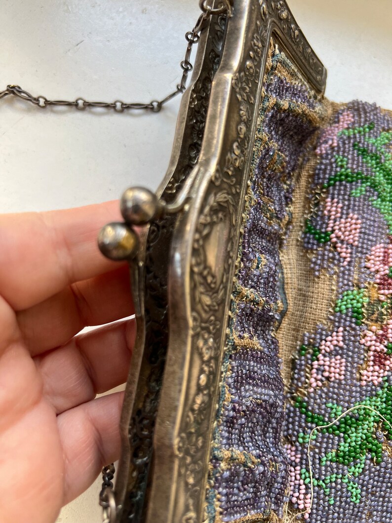 May include: A vintage beaded purse with a silver frame and a chain strap. The purse is decorated with a floral pattern in shades of purple, pink, and green.