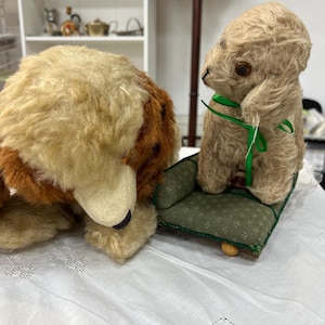 Può includere: Due giocattoli di peluche vintage: un cane e un coniglio. Il cane è marrone e marrone chiaro, con un naso nero. Il coniglio è marrone chiaro, con un nastro verde intorno al collo, seduto su un piccolo cuscino verde.