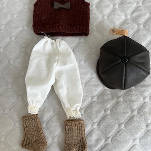Puede incluir: Un conjunto de ropa de muñeca que incluye un chaleco de punto marrón con lazo, pantalones blancos con ribete de encaje, calcetines de punto marrones y una gorra marrón oscuro. El conjunto está dispuesto sobre una superficie clara y texturizada.