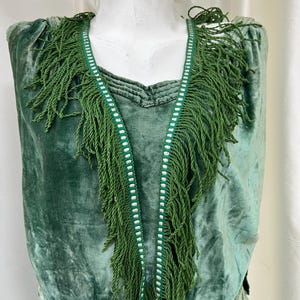 May include: A vintage emerald green velvet dress with a fringed collar. The dress features a V-neckline and is adorned with green fringe and white trim. The bottom of the dress has embroidered floral details with pink accents.