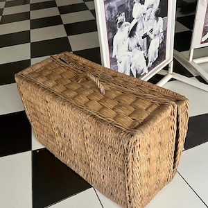 May include: A rectangular, woven wicker basket with a handle. The basket is light brown and has a tightly woven pattern. The basket is sitting on a black and white checkered floor. A framed black and white photo is in the background.
