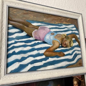 May include: Oil painting in a white frame shows a person lying on a blue and white striped towel. The person wears a light blue top and pink shorts. The artwork is a beach scene.