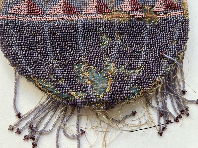 May include: A close-up of a beaded purse with a fringe of purple beads. The purse is made of a dark green fabric with a floral pattern.