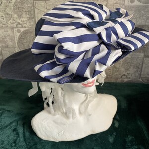 May include: A navy blue sun hat with a wide brim and a large bow made of white and navy blue striped fabric. The hat is displayed on a white bust, highlighting its design and style.